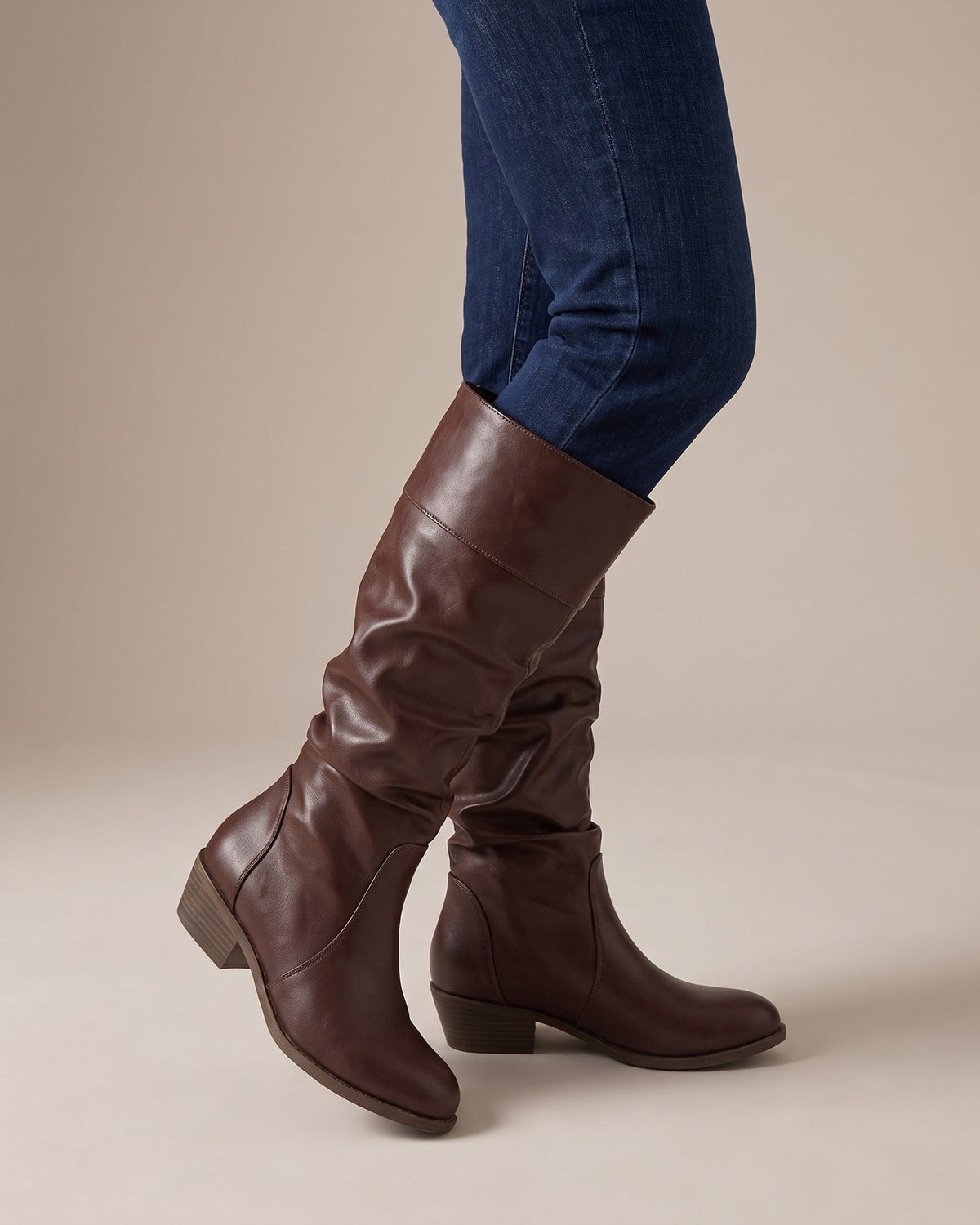 Brown knee-high riding boots in smooth leather-look material with a slouchy shaft, cuffed top, and low stacked heel.