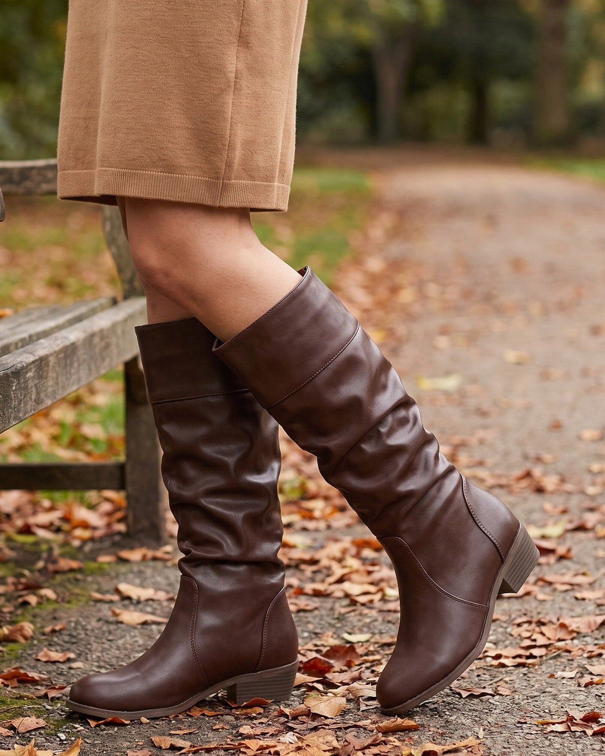 Brown knee-high riding boots with a slouchy shaft, cuffed top, and low stacked heel worn with a tan sweater dress.