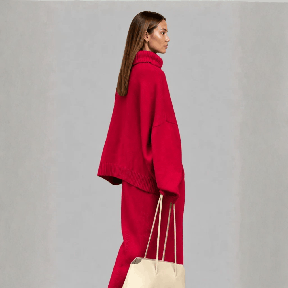 Side profile view of a model wearing the bright red oversized turtleneck sweater and skirt set, holding a cream tote bag.