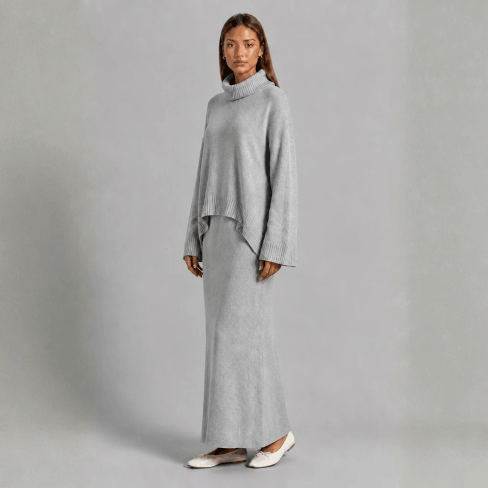 Three-quarter front view of a model wearing the oversized turtleneck sweater and maxi skirt set in light gray, styled with white flats.   