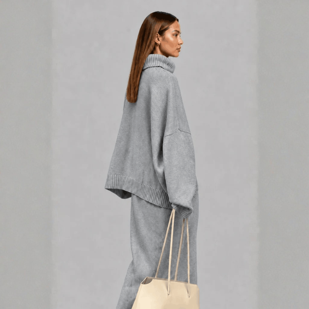 Side profile view of a model wearing the light gray oversized turtleneck sweater and maxi skirt set, holding a cream tote bag.