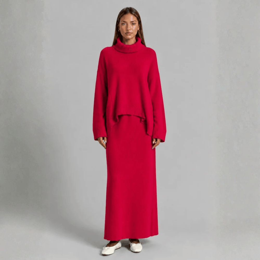 Full-length front view of a model wearing a bright red knit two-piece set featuring an oversized turtleneck sweater with dropped shoulders and a matching column maxi skirt, styled with white ballet flats.
