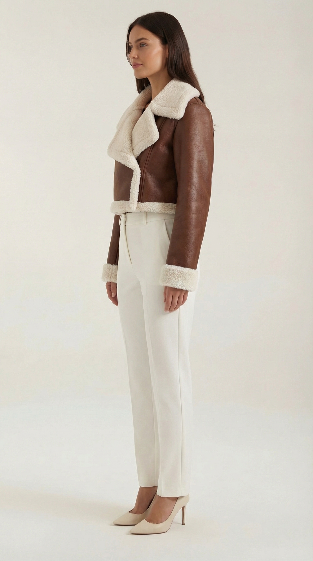 Brown leather aviator jacket with a cropped fit, plush cream shearling trim, and zip-up front worn with white trousers.
