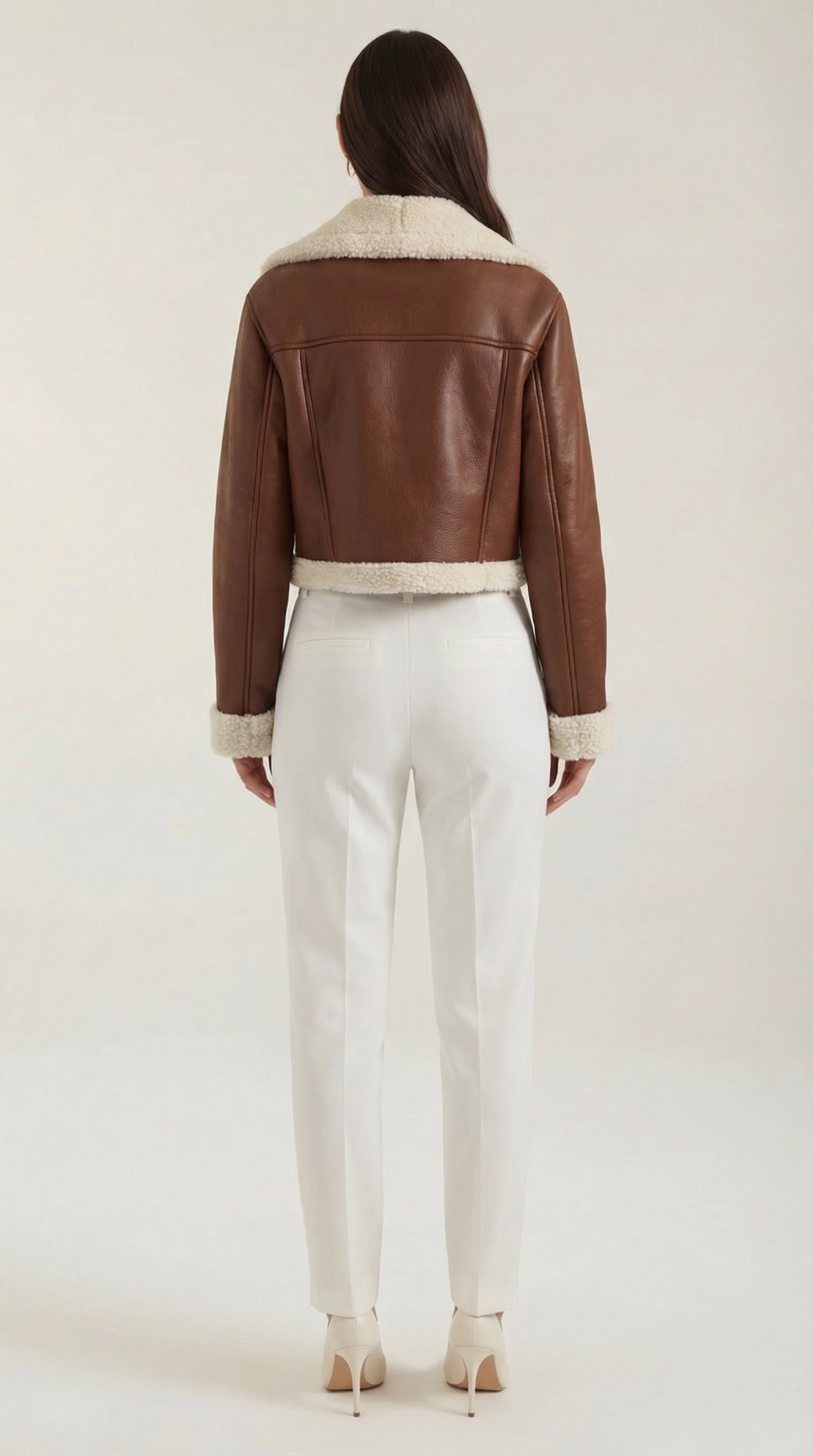 Brown leather aviator jacket with a cropped fit, cream shearling trim, and fleece lining, worn with white trousers.