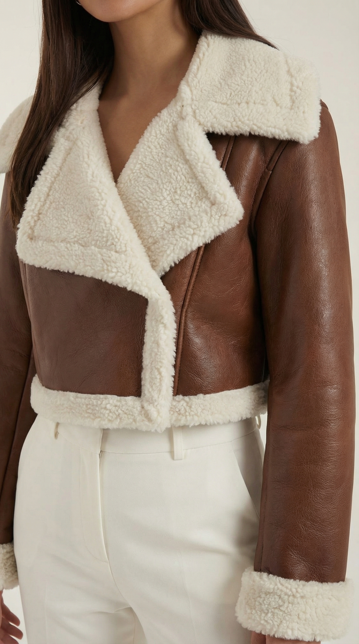Brown leather aviator jacket with a cropped fit, plush cream shearling trim, and zip-up front worn over white trousers.