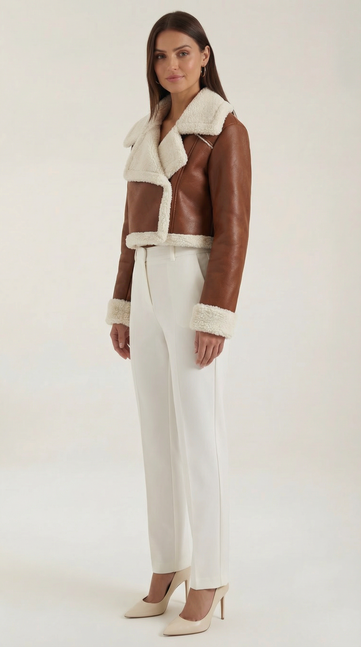 Brown leather aviator jacket with a cropped fit, plush shearling trim, and fleece-lined collar worn with white trousers.