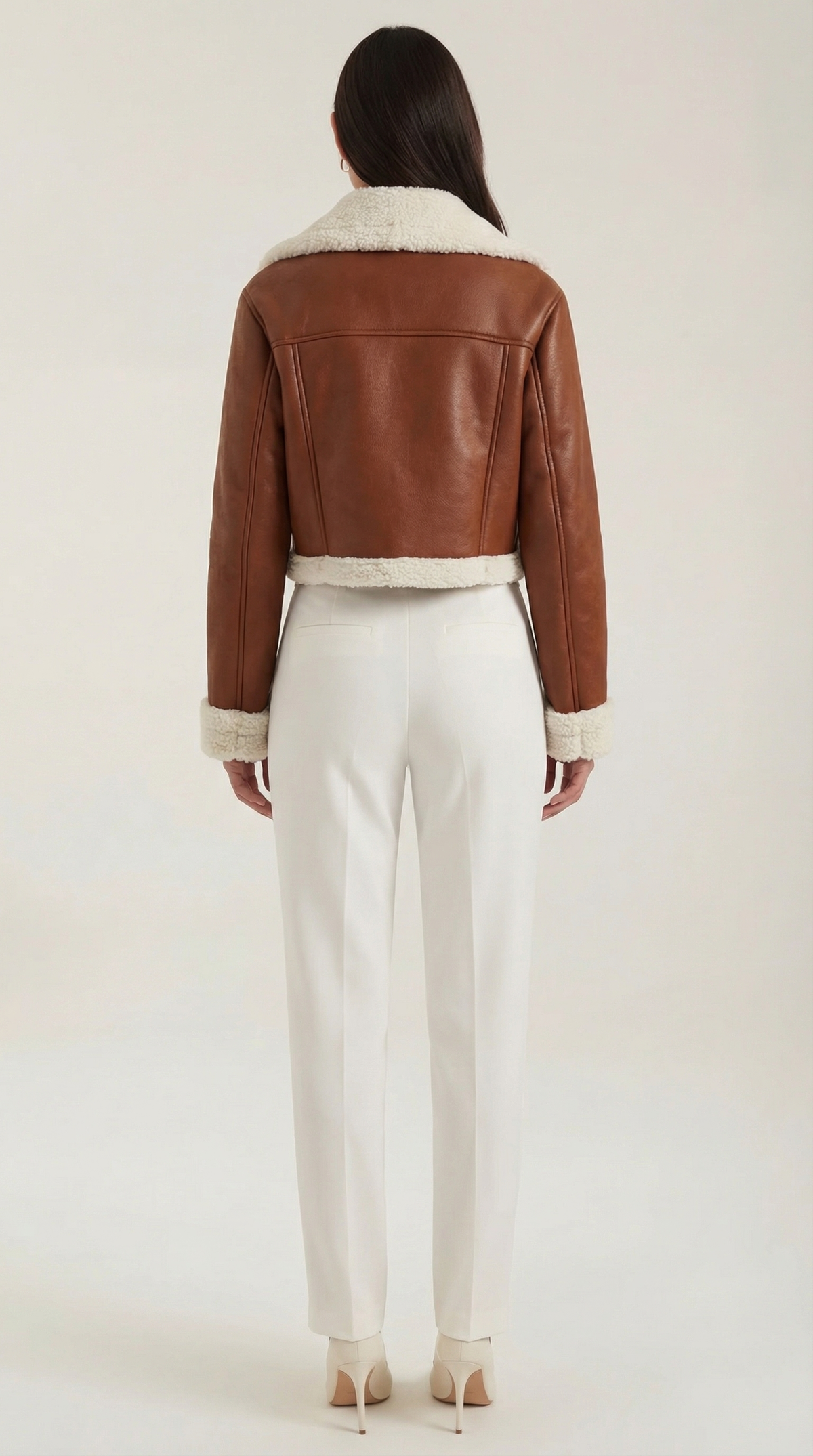 Brown leather aviator jacket with a cropped fit, white shearling trim, and fleece lining worn with white trousers.