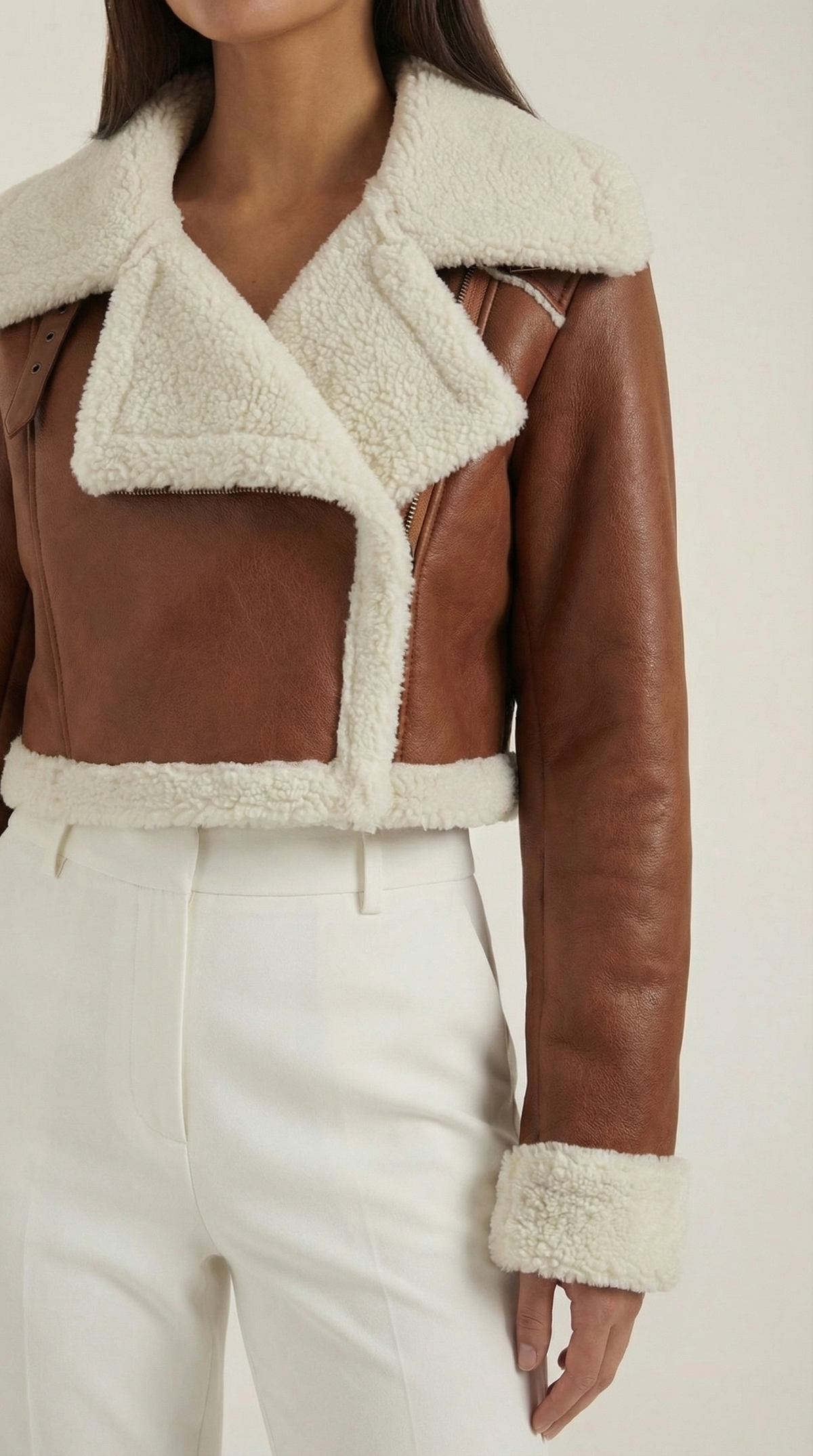 Brown leather aviator jacket with a cropped fit, plush cream shearling trim, and zip-up front worn with white trousers.