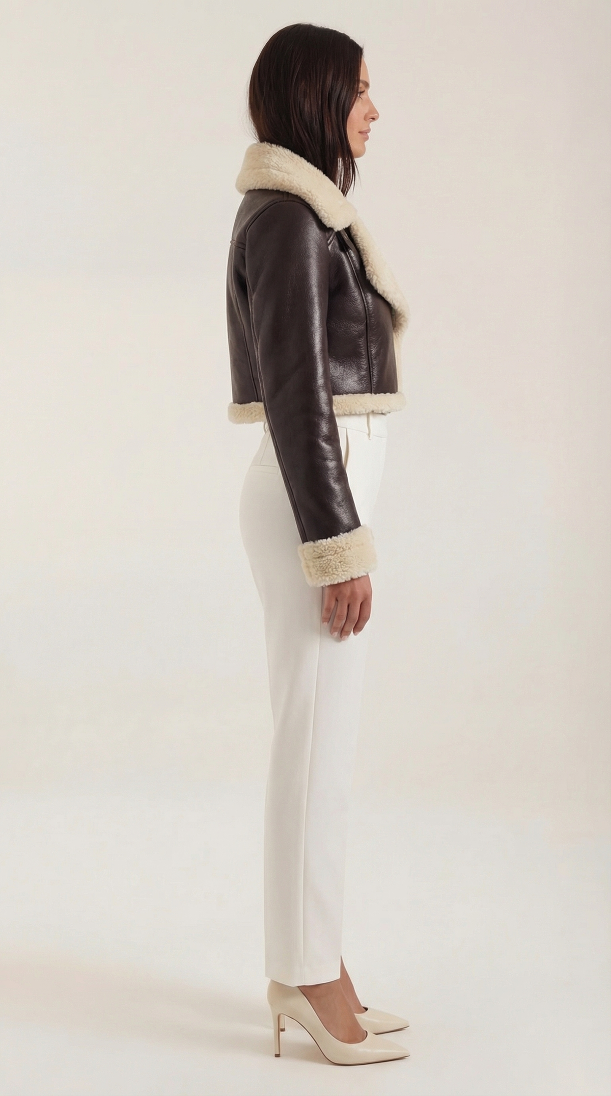 Brown leather aviator jacket with a cropped fit, cream shearling trim, and fleece lining worn with white trousers.