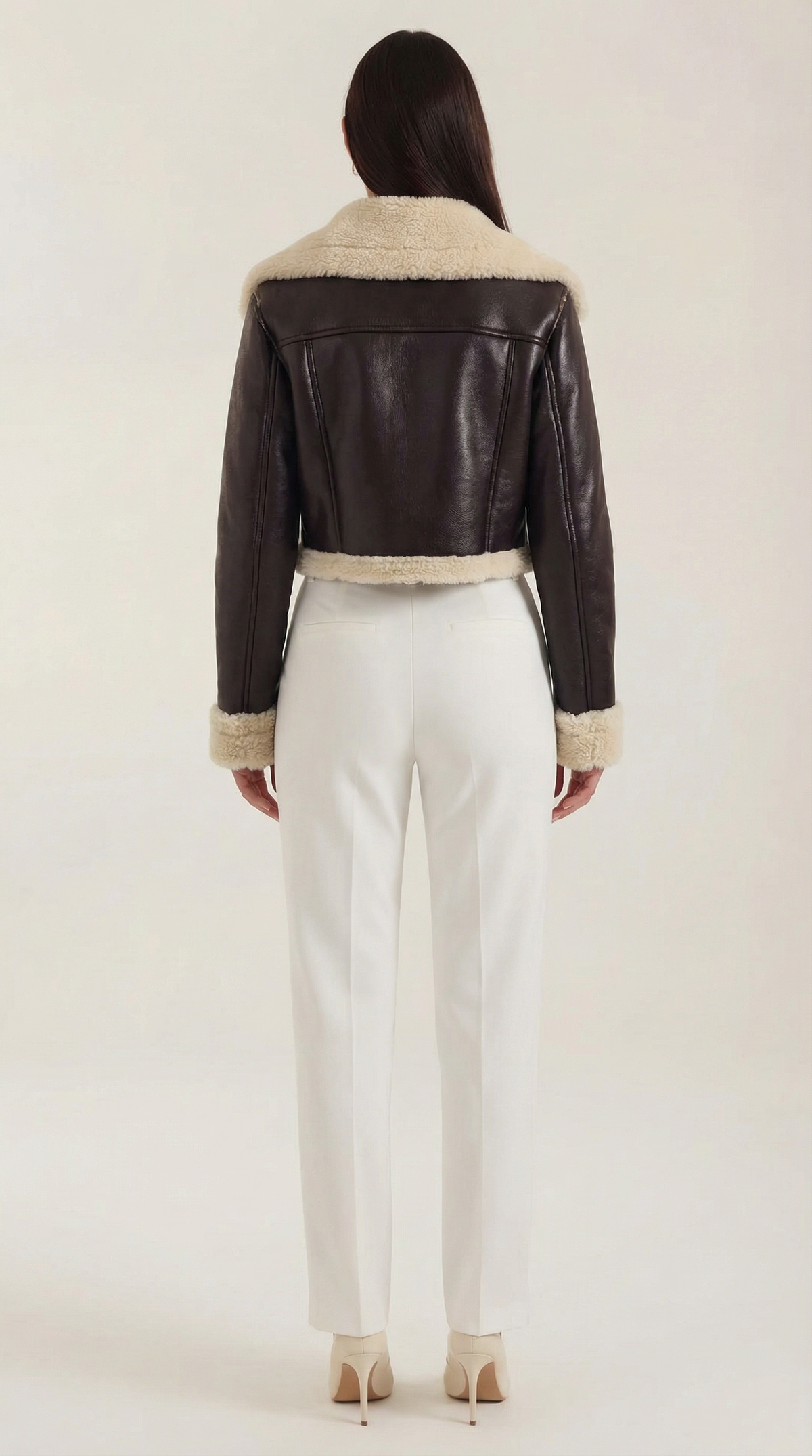 Brown leather aviator jacket with cream shearling trim, cropped fit, and fleece lining worn with white trousers.