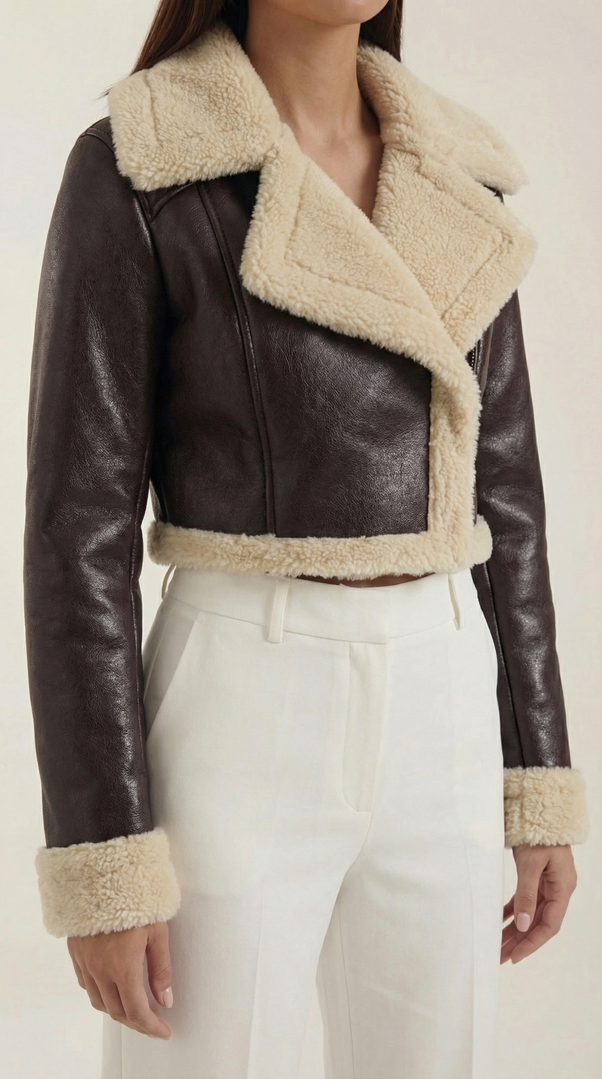 Brown leather aviator jacket with a cropped fit, cream shearling trim, and zip-up front worn with white trousers.