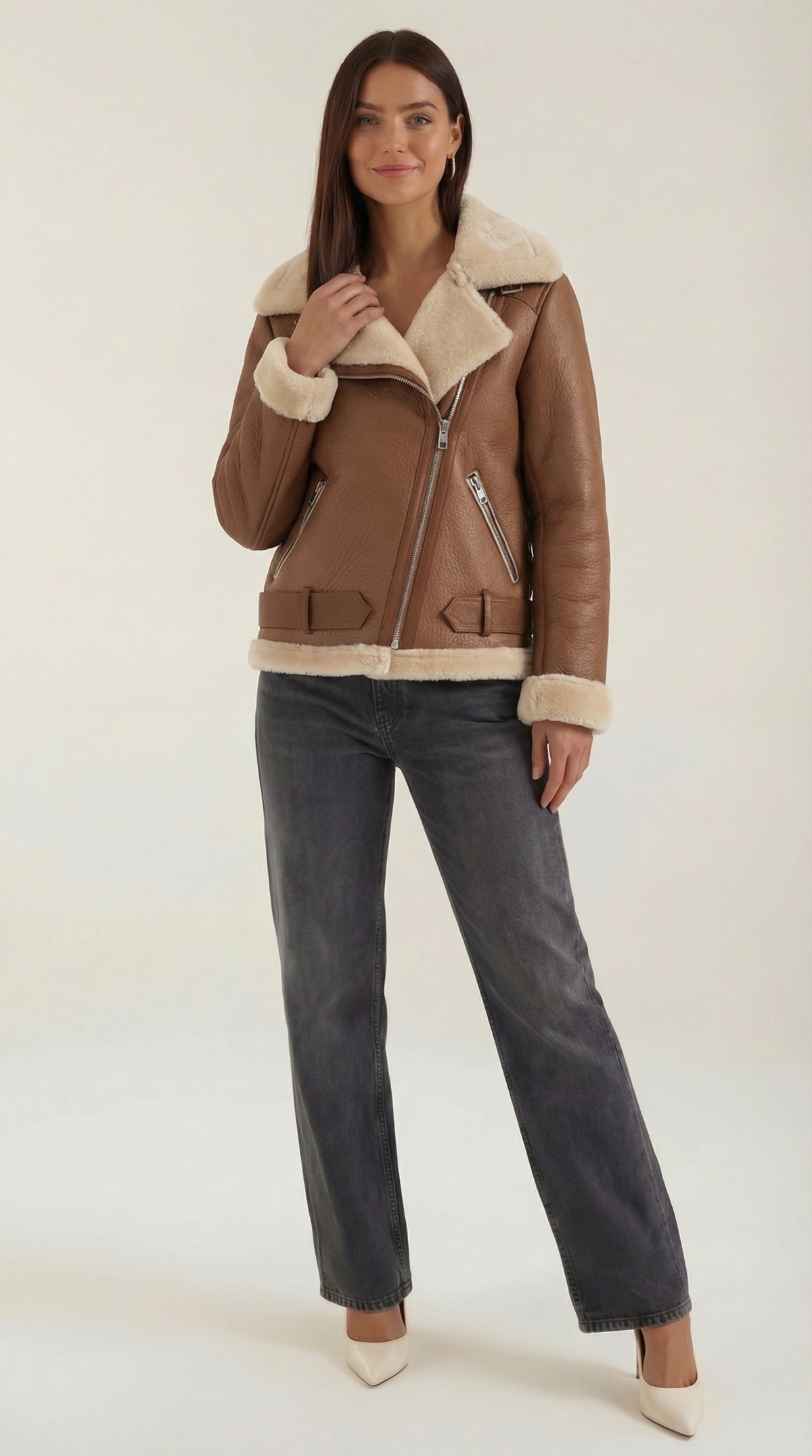 Brown leather aviator jacket with cream shearling trim, diagonal zip front, and belted hem worn with grey jeans.