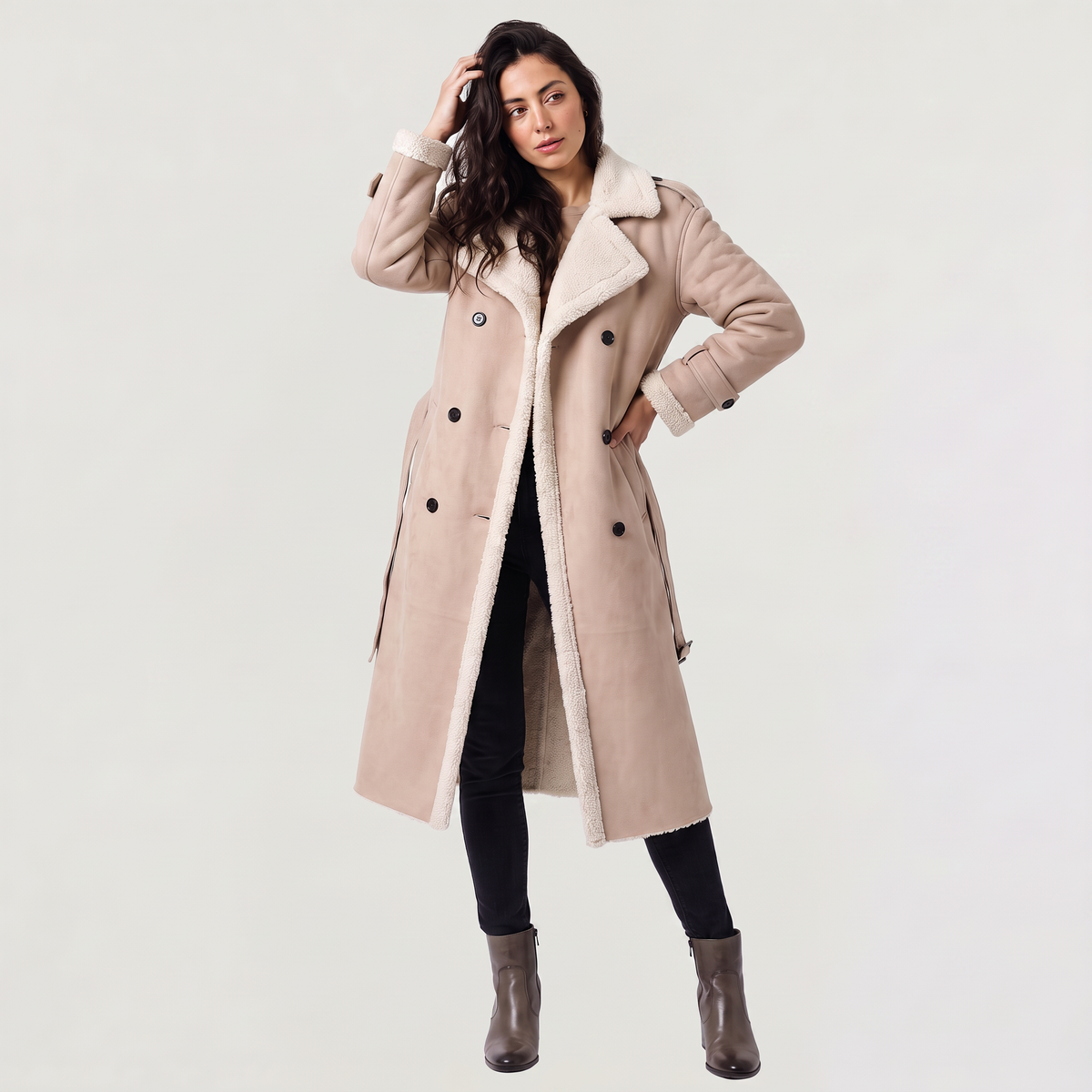 A woman wearing a apricot longline leather aviator coat with borg trim and tie waist, posing with one hand on her waist