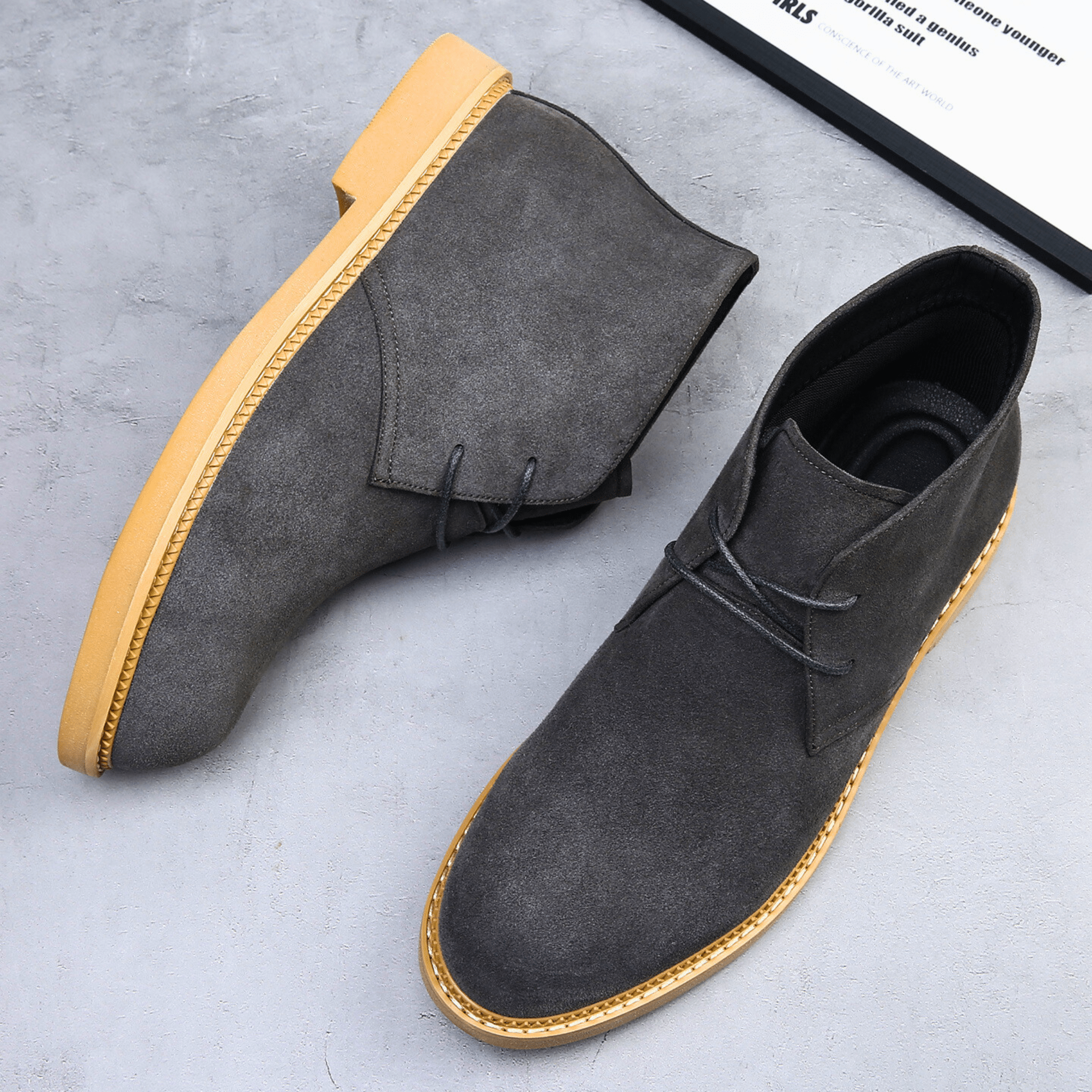 Charcoal grey suede-look desert boots with a plain toe, two-eyelet lacing, and a contrasting tan stacked sole on grey stone.