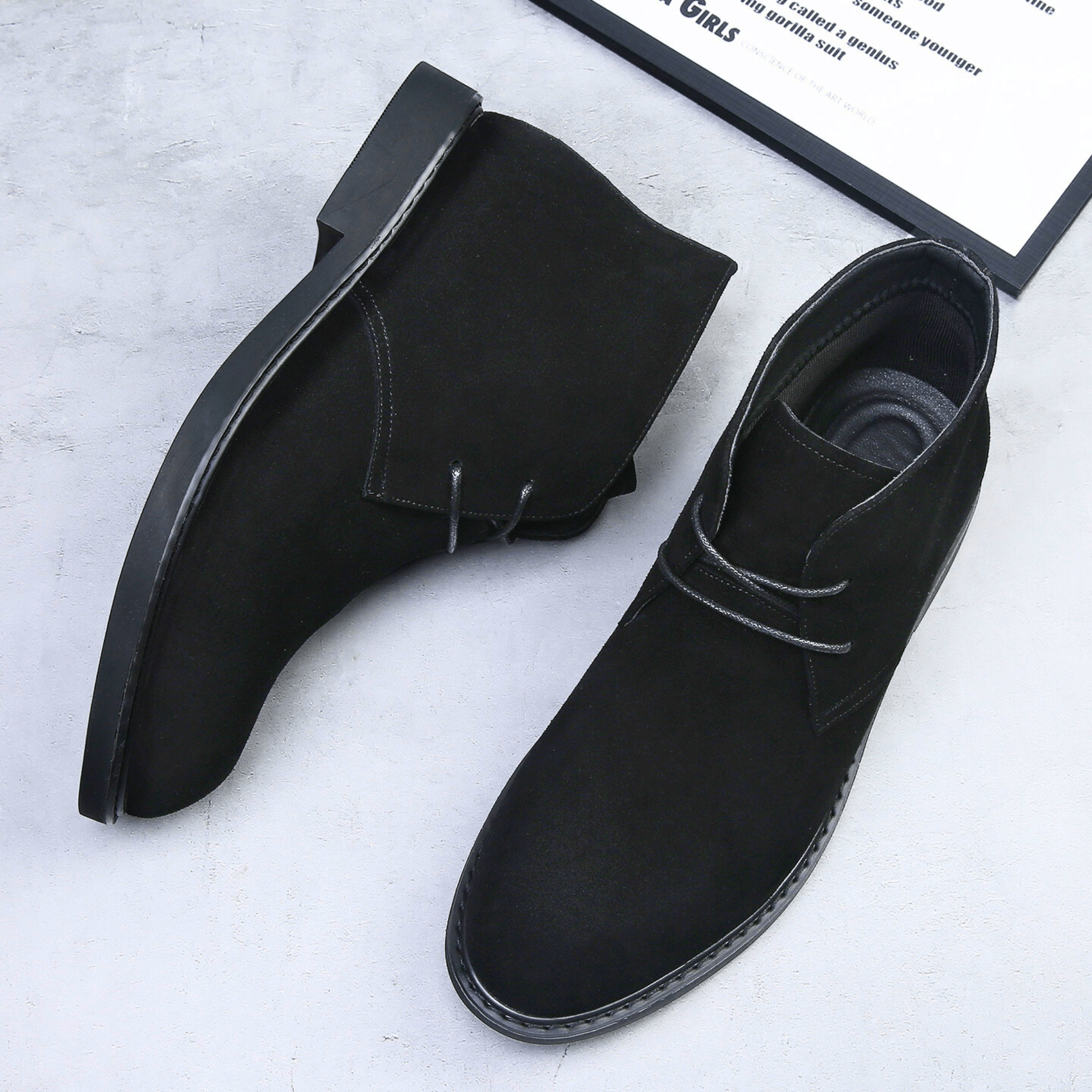 Black suede-look desert boots with a plain toe, two-eyelet lacing, and a low stacked heel on a grey surface.