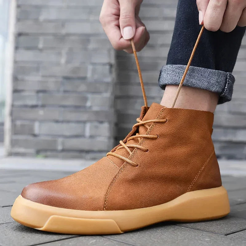Tan leather chukka boots with a rounded toe and chunky rubber sole, shown being laced up with dark cuffed jeans.