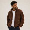 A dark-haired man wearing a dark brown men's full-zip fleece jacket with a high stand collar, chest yoke seam, and zippered side pockets, styled open over a white t-shirt and dark trousers.