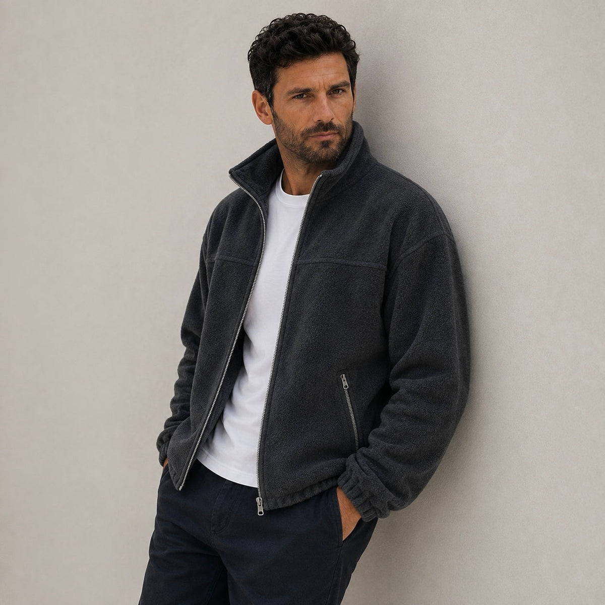 A man leaning against a neutral background wearing a dark charcoal gray zip-front fleece jacket with zippered hand pockets.