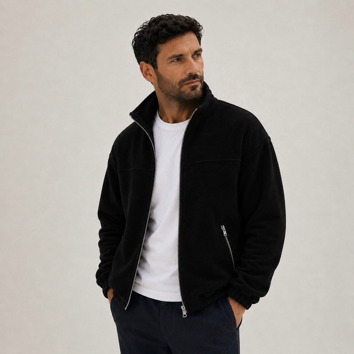 A man wearing a black full-zip fleece jacket with a high collar, styled over a white t-shirt and dark trousers.