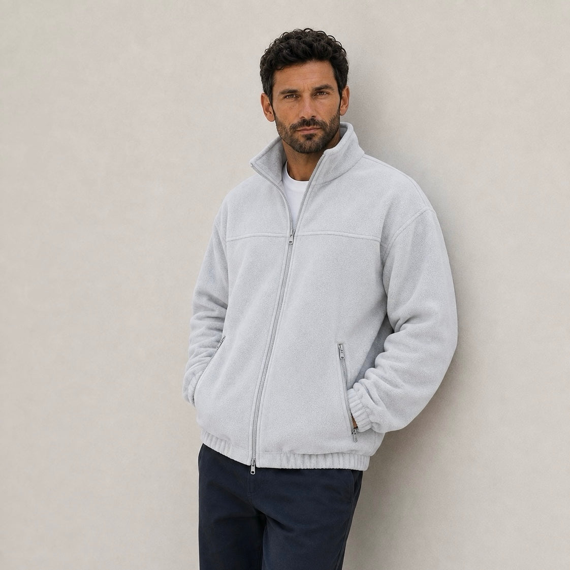 A man leaning against a wall wearing a light gray fleece jacket zipped up to the high collar, worn over dark trousers.