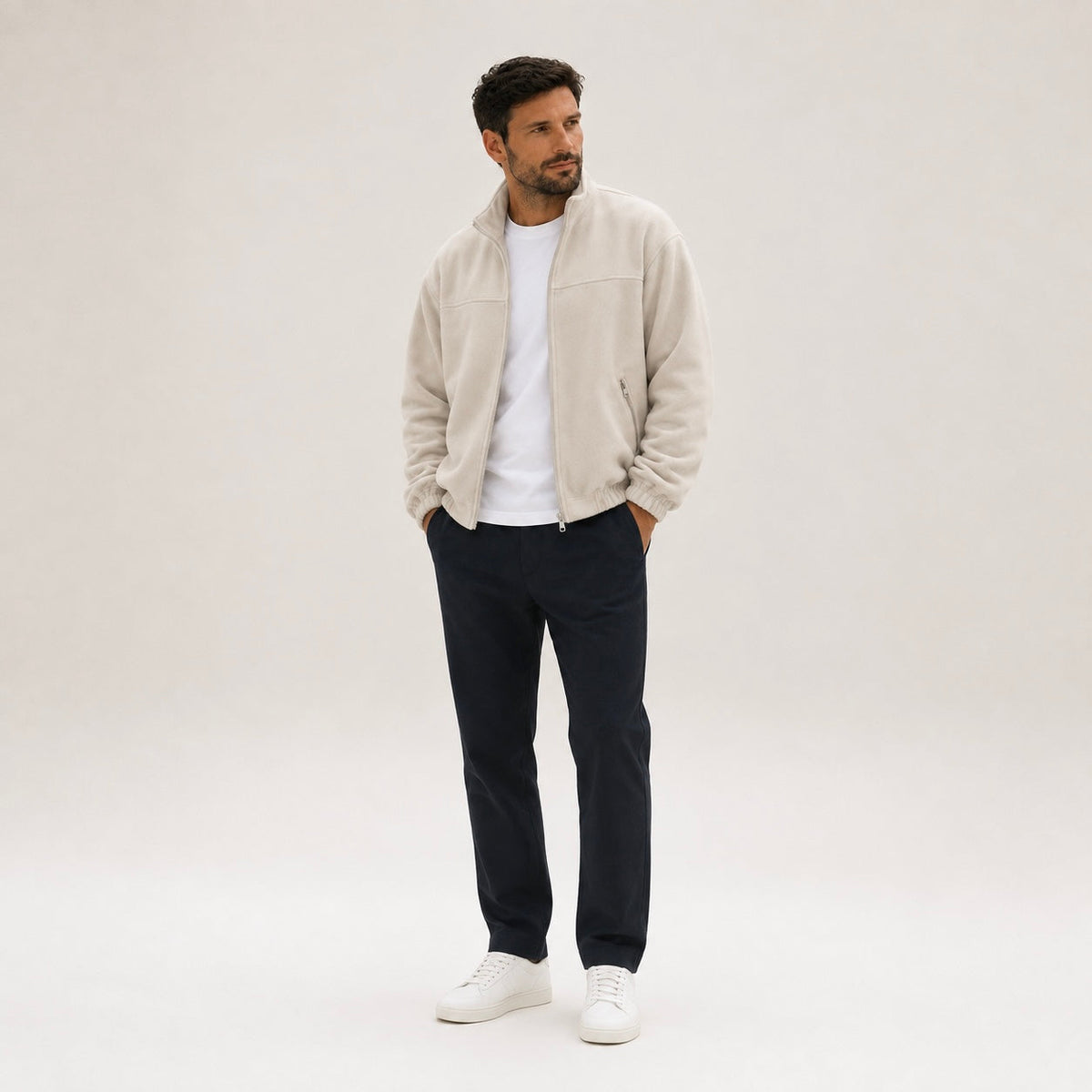 Full-length view of a man wearing an cream full-zip fleece jacket, dark navy trousers, and white sneakers.