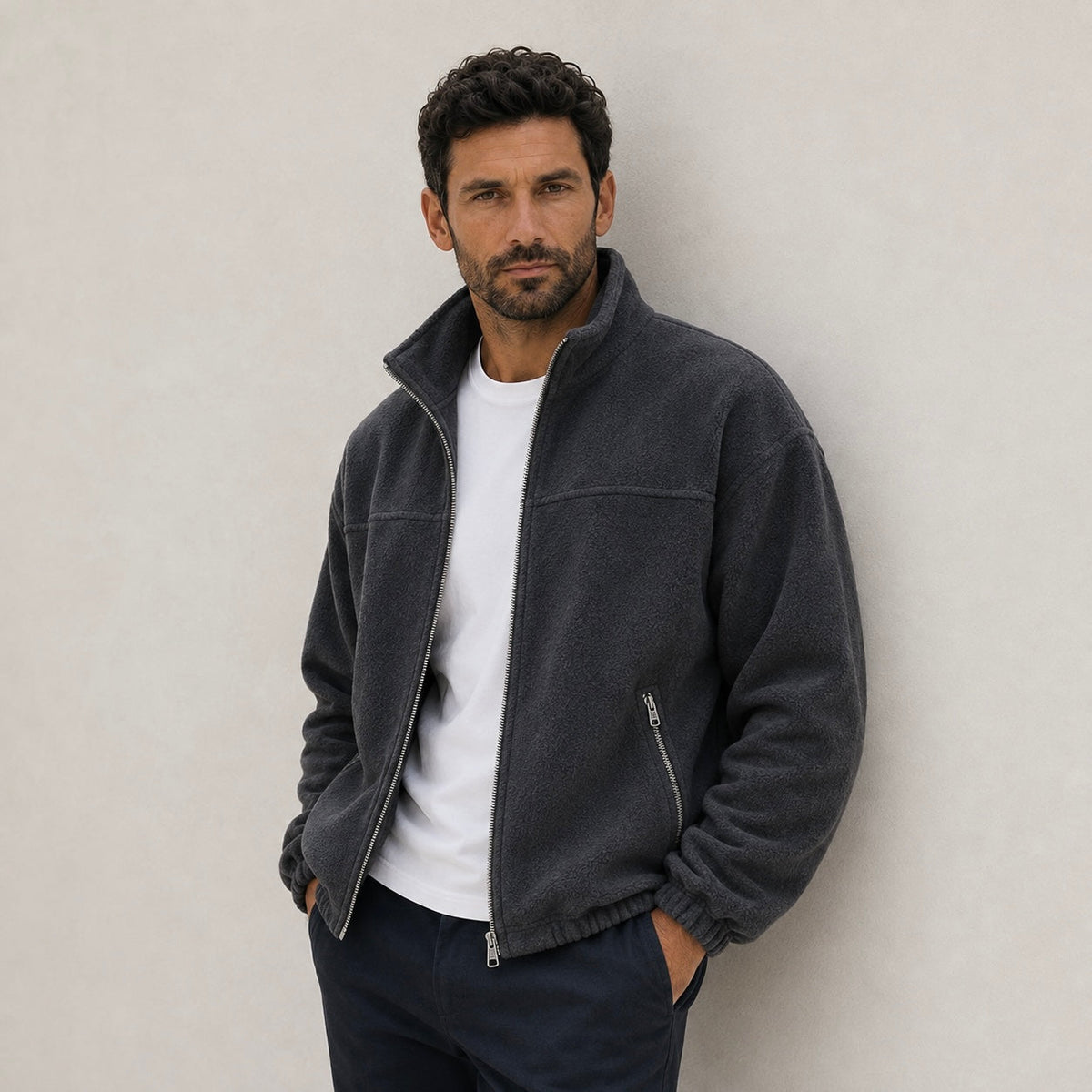 A man wearing a dark charcoal gray full-zip fleece jacket with a stand-up collar, layered open over a plain white crewneck shirt and dark pants.