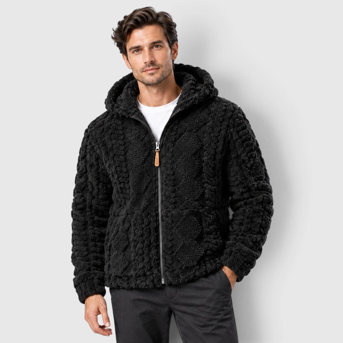 A man wearing a black cable-textured hooded fleece jacket zipped up over a white t-shirt, paired with dark pants.