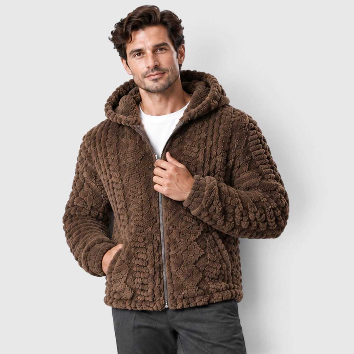 A dark-haired man wearing a dark brown hooded fleece jacket with an embossed cable-texture pattern and full front zipper, styled over a white t-shirt and dark trousers.