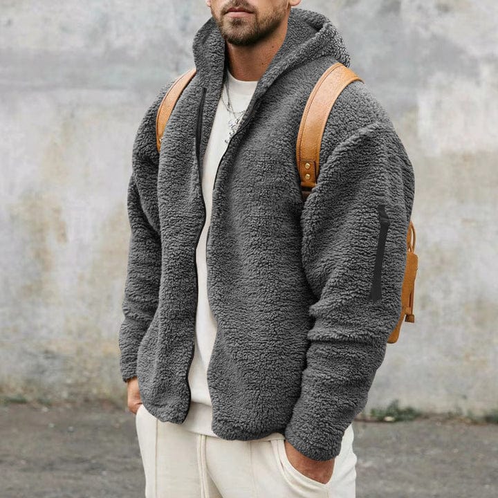 A man wearing a loose-fit dark gray hooded teddy fleece jacket with a full-zip front, styled with a white shirt, white pants, and a leather backpack.