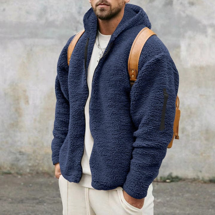 A man wearing a navy blue loose-fit hooded teddy fleece jacket worn open over a white t-shirt, white pants, and a tan backpack.