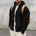 A man wearing a loose-fit black hooded teddy fleece jacket open over a white t-shirt and white pants, carrying a light brown backpack.