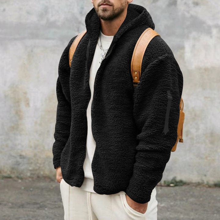 A man wearing a loose-fit black hooded teddy fleece jacket open over a white t-shirt and white pants, carrying a light brown backpack.