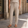 Beige merino wool pants with a drawstring waist and relaxed tapered fit, worn with leather sandals on a stone street.