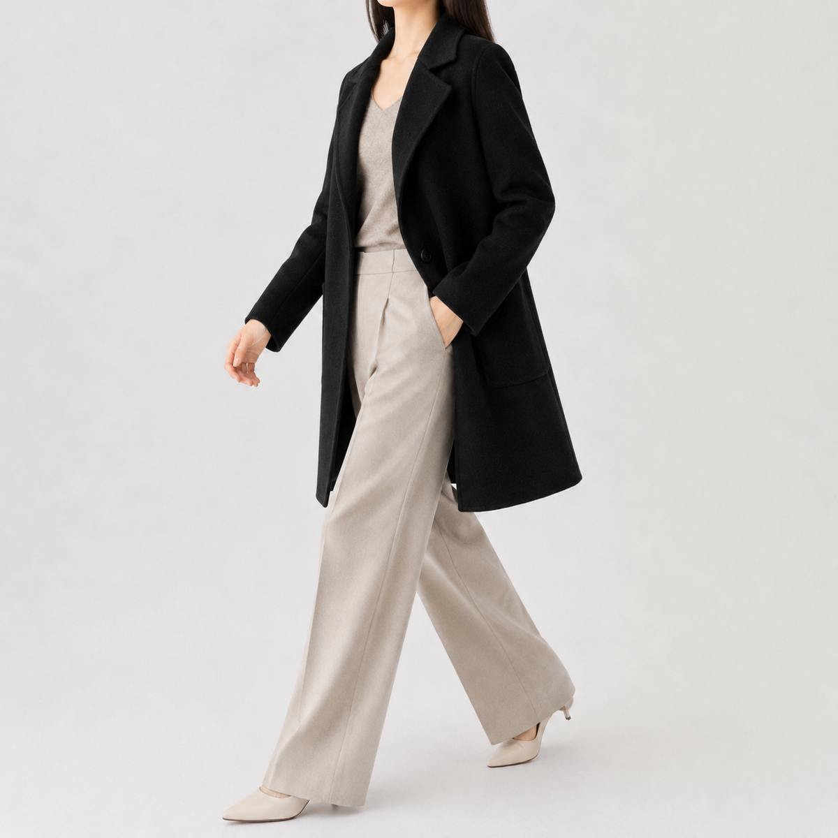 Black mid-length wool coat with a notch lapel, single-button closure, and patch pockets, worn over neutral trousers.