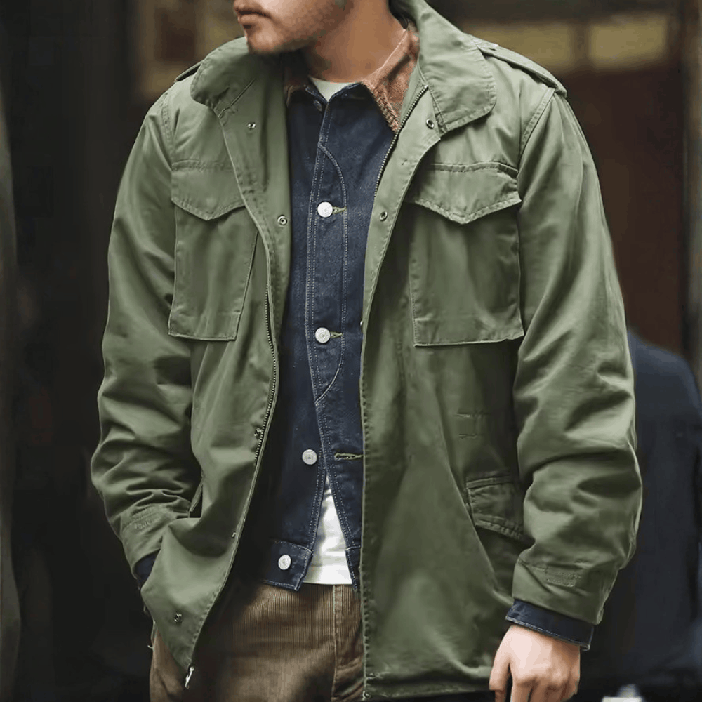 Three-quarter front view of a model wearing the olive green military field jacket layered over a denim shirt outdoors.