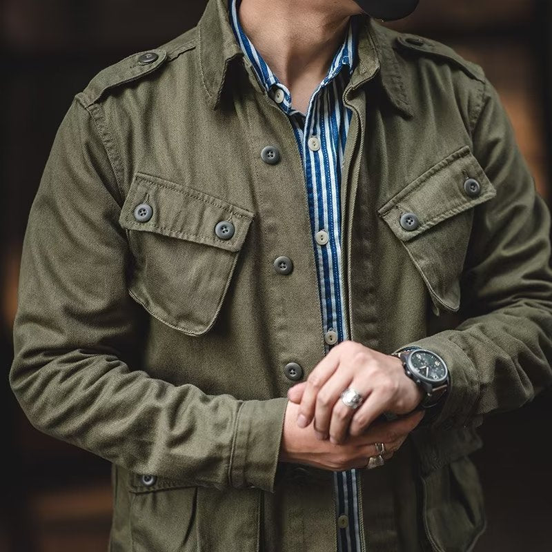Close-up view of the upper torso of the olive green military utility jacket, showing the buttoned chest cargo pockets, shoulder epaulets, button placket, and wristwatch detail.