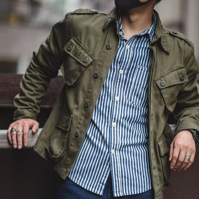 Model leaning against a balcony rail wearing the open olive green military-style utility jacket over a blue and white striped button-up shirt.