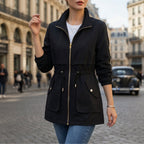 Black cotton blend utility jacket with a zip-front, drawstring waist, and flap pockets, worn on a city street.