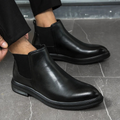 Black leather Chelsea boots with a round toe, elastic side panels, and a chunky treaded lug sole on a grey tiled floor.