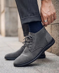 Grey suede chukka boots with a plain toe, lace-up front, heel pull tab, and a black molded sole with visible moc stitching.