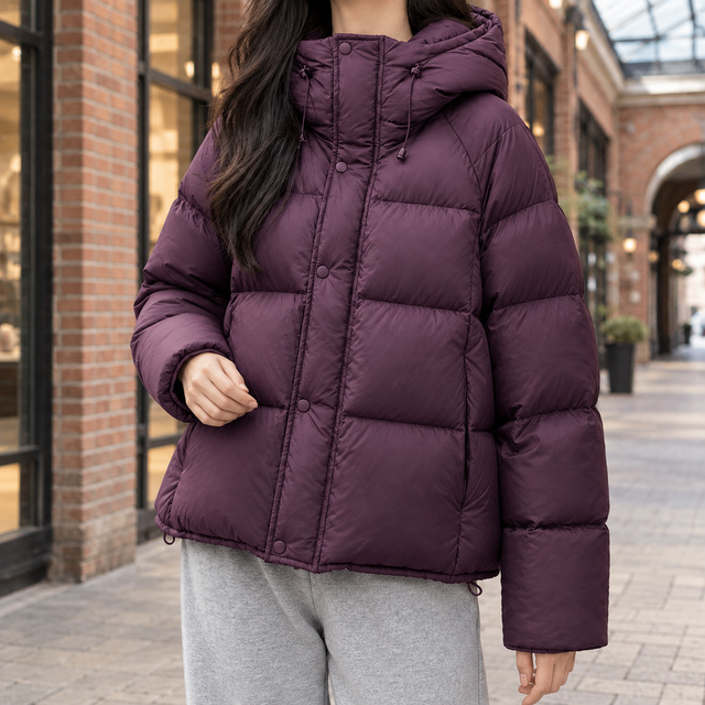 Deep purple oversized puffer jacket with wide box quilting, snap-over zip front, and adjustable hood on a city street.