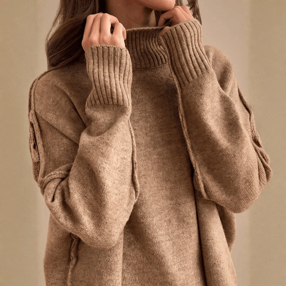 Close-up view of a model adjusting the ribbed turtleneck collar on the beige oversized knit sweater, highlighting the dropped shoulder seam details.