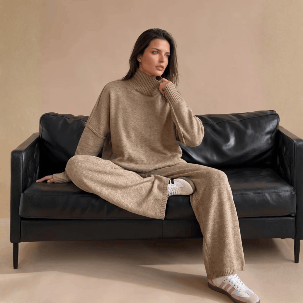 odel sitting cross-legged on a black leather sofa wearing the beige oversized turtleneck knit lounge set.