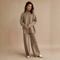 Full-length front view of a model wearing a beige oversized turtleneck knit lounge set featuring a high ribbed collar, dropped shoulder seams, side slits, and wide-leg trousers, styled with white sneakers.