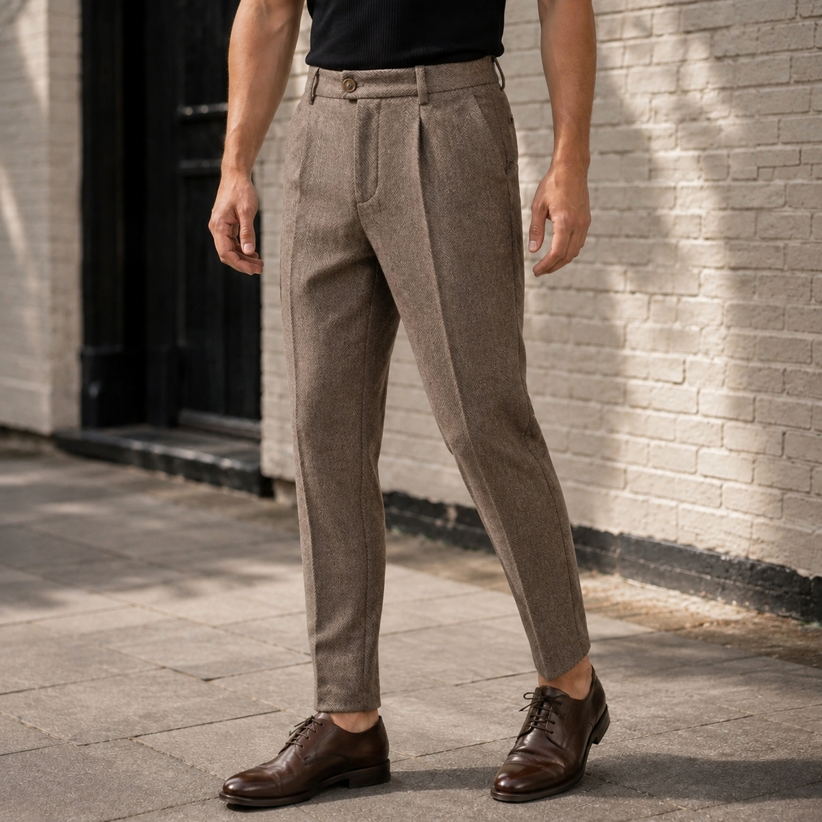 Full-length side-angle shot of a man wearing straight-leg pleated herringbone wool dress pants and brown leather shoes, posing outdoors near a brick building.