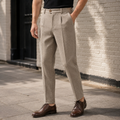 Front three-quarter view of a man wearing light beige-brown pleated herringbone wool pants with a buttoned waistband extension, straight legs, and brown leather derby shoes, paired with a black t-shirt outdoors against a brick wall.   