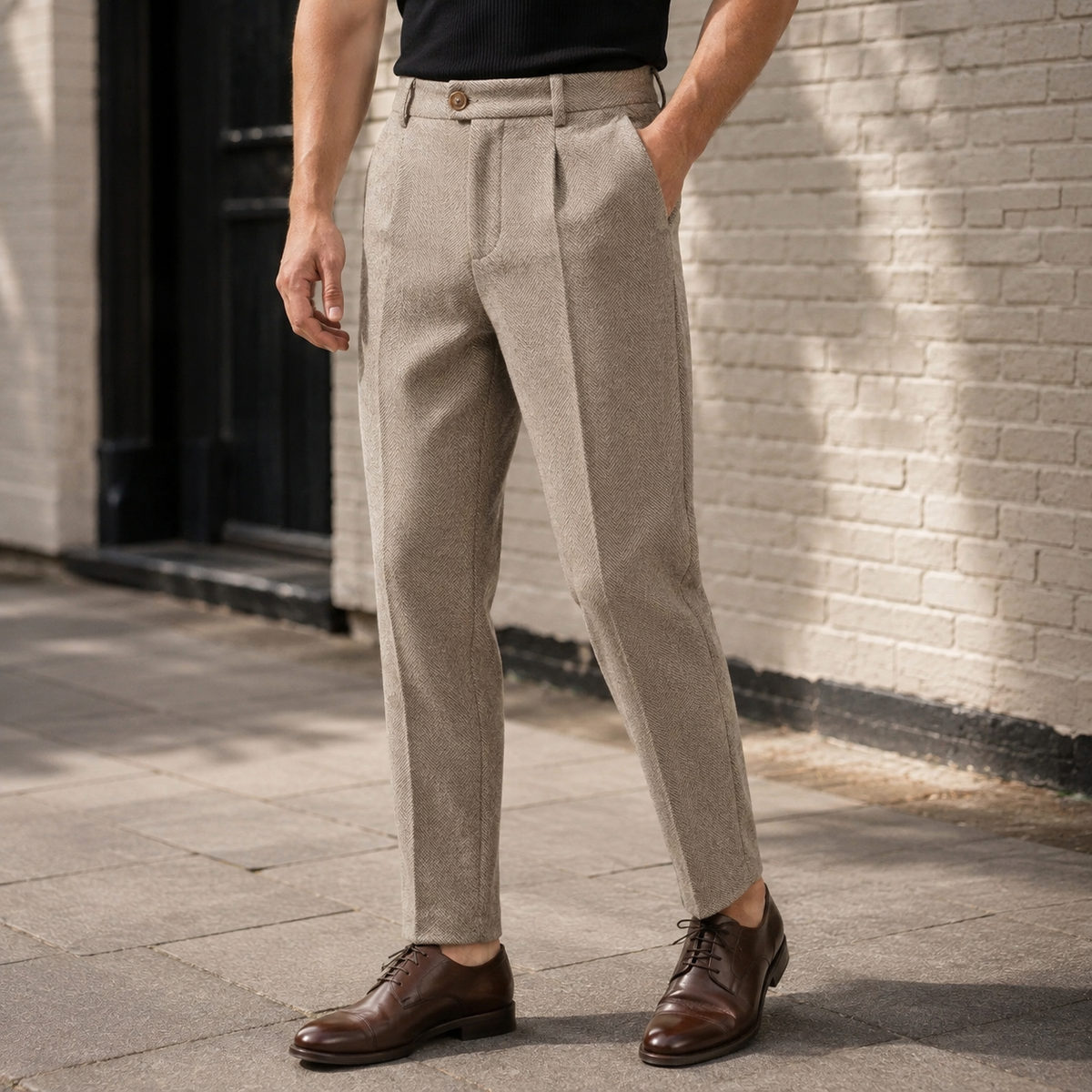 Front three-quarter view of a man wearing light beige-brown pleated herringbone wool pants with a buttoned waistband extension, straight legs, and brown leather derby shoes, paired with a black t-shirt outdoors against a brick wall.   