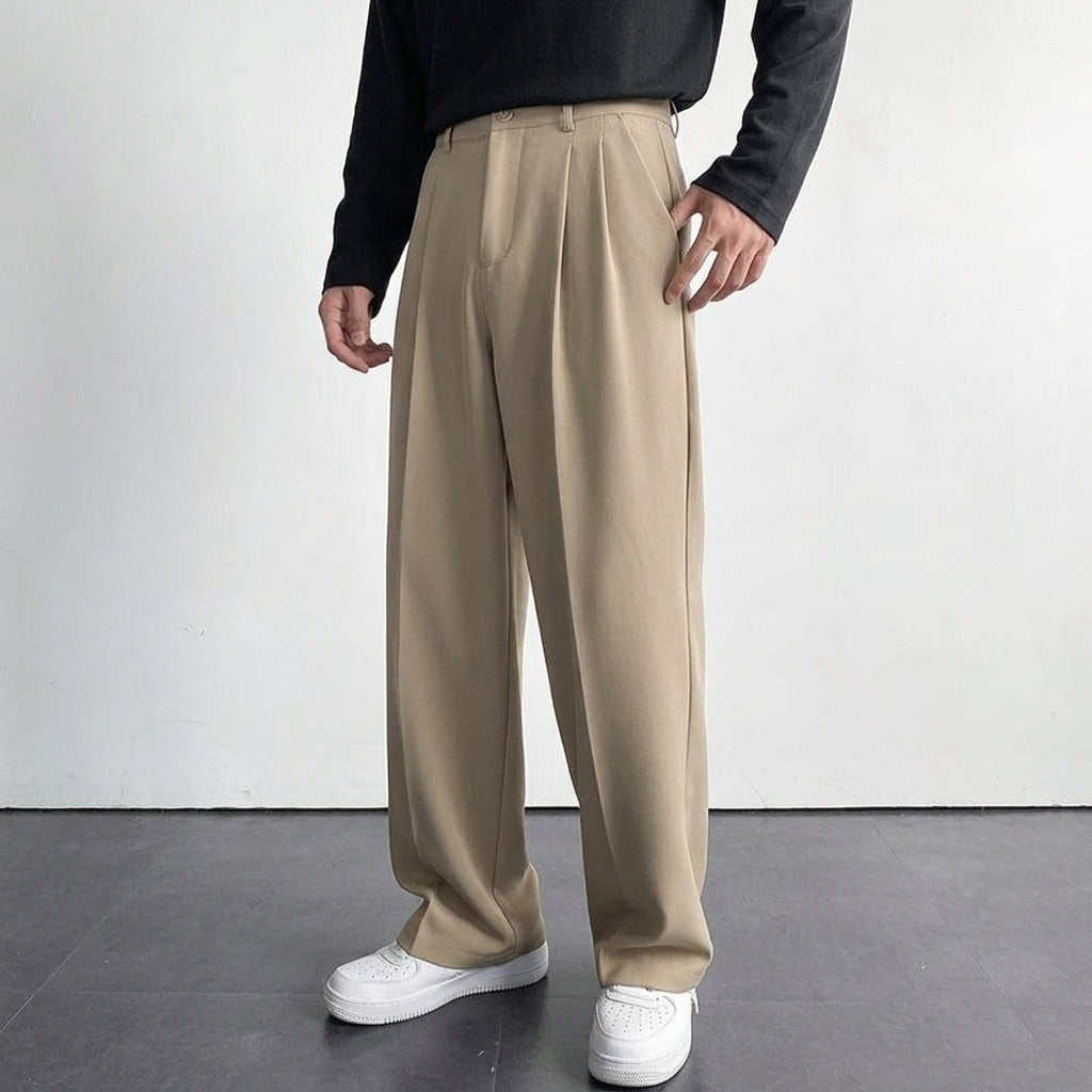 Beige pleated wool trousers with a relaxed, full-length drape and tailored front detailing worn with a black top.