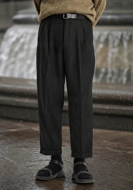 Front view of charcoal black pleated wool tweed dress pants with a cuffed hem, styled with a beige sweater, black belt, black socks, and strap sandals outdoors.