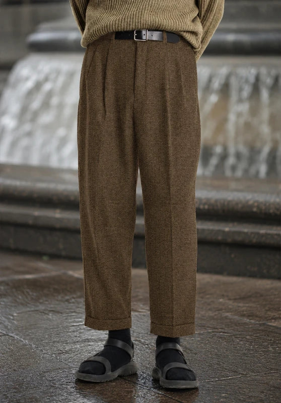 Front view of khaki pleated wool tweed dress pants with cuffed hems, paired with a tucked-in tan sweater, black belt, black socks, and sandals.  